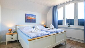 A bedroom with a bed, bedside tables, a painting, a window with curtains, and a radiator. - At Home Comfortable Holiday Residence (Borkum)