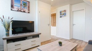 A living room with a flat-screen TV on a wooden stand, a coffee table, and a door leading to another room. - At Home Comfortable Holiday Residence (Borkum)