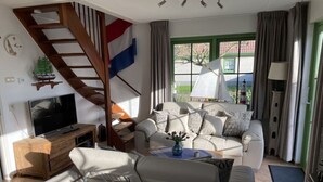 A living room with a sofa, a TV, a wooden cabinet, a flag, and a window with curtains. - "sina" Comfortable Holiday Residence (Julianadorp)