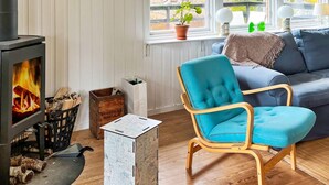 A cozy living room with a wood-burning stove, a blue armchair, a sofa, and a view of trees through the window. - 4 Star Holiday Home in Vato (Vätö)