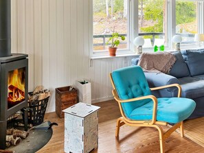 A cozy living room with a wood-burning stove, a blue armchair, a sofa, and a view of trees through the window. - 4 Star Holiday Home in Vato (Vätö)