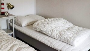 Un lit simple avec un matelas et des oreillers blancs matelassés, une table de chevet avec une lampe et une horloge fixée au mur.