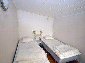 A small, single-room accommodation with two single beds, a bedside table with lamps, and a plain white wall.