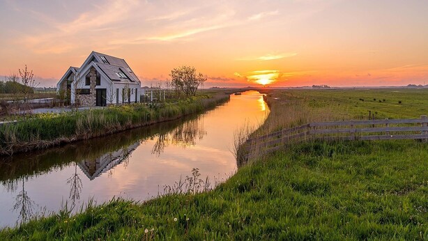 A riverside house with a stone facade and a sloped roof, surrounded by lush greenery and a calm river. - Energy-neutral Villa for 2 People (Oudeschild)