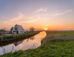 A riverside house with a stone facade and a sloped roof, surrounded by lush greenery and a calm river. - Energy-neutral Villa for 2 People (Oudeschild)