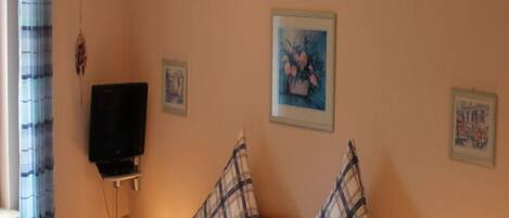 Una sala de estar con un televisor sobre un mueble bajo, una mesita de noche con una lámpara y varias fotografías enmarcadas en la pared.
