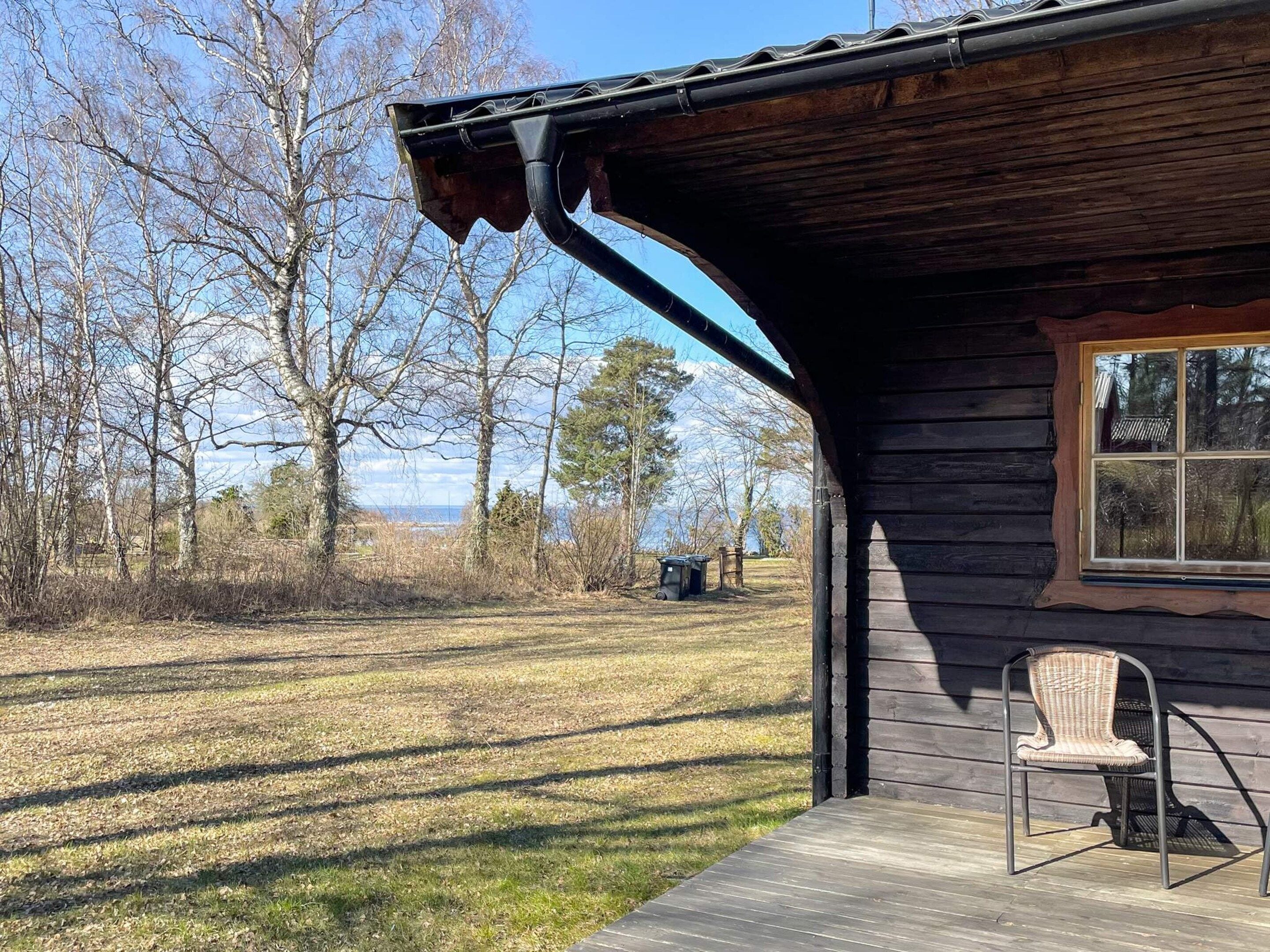 Una cabaña de madera con un porche, una silla y vistas a árboles y un cuerpo de agua.