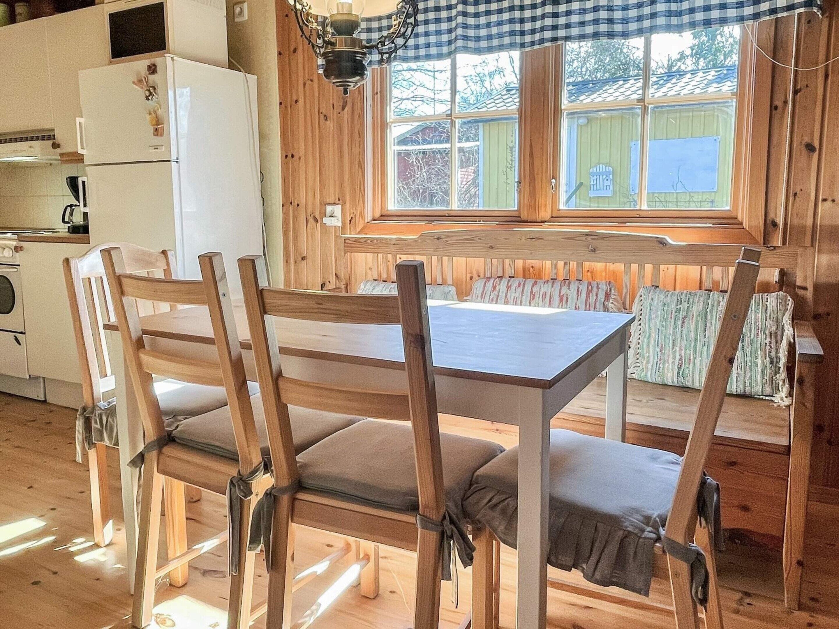 A wooden dining table with chairs, a kitchen area with a refrigerator, and a window with a view of the outside.