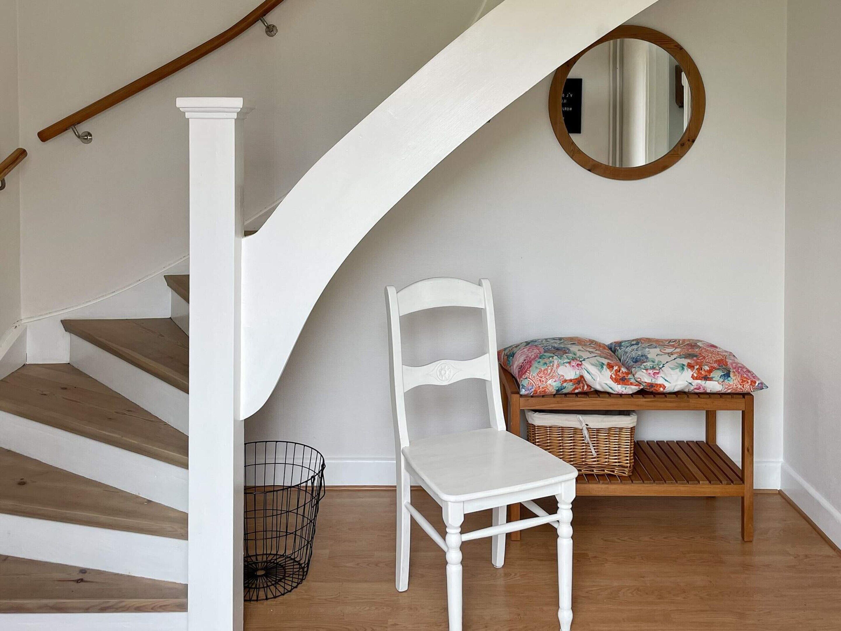 Eine Treppe mit Holzbalken und weißen Geländern, ein weißer Stuhl, eine Bank mit Blumenkissen und ein runder Spiegel an der Wand.
