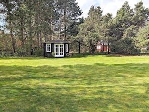 A small house with a white door and windows, surrounded by a grassy lawn and trees. - 4 Person Holiday Home in Rudkobing (Rudkobing)
