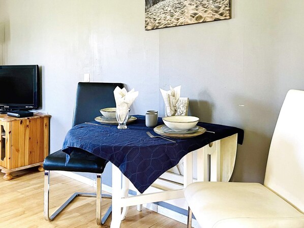 Un comedor con una mesa dispuesta para dos, un televisor sobre un mueble de madera y una silla blanca.