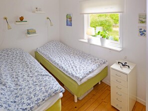 Une chambre avec deux lits simples, une fenêtre donnant sur un espace verdoyant, une table de chevet avec une lampe et une commode avec des tiroirs.