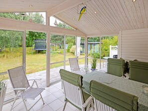 Une terrasse couverte avec une table et des chaises, un oiseau suspendu au plafond et une vue sur un jardin avec un portique.