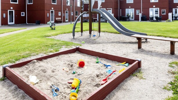 A playground with a slide, sandpit, and play structures, surrounded by red buildings. - 6 Person Holiday Home in Hadsund (Hadsund)