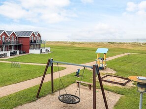 A playground with swings and a seesaw, a red building with a balcony, and a sandy area in the background. - 6 Person Holiday Home in Hadsund (Hadsund)