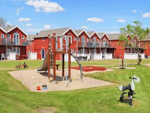 A playground with a slide and climbing structures, surrounded by a grassy area and red buildings with balconies in the background.