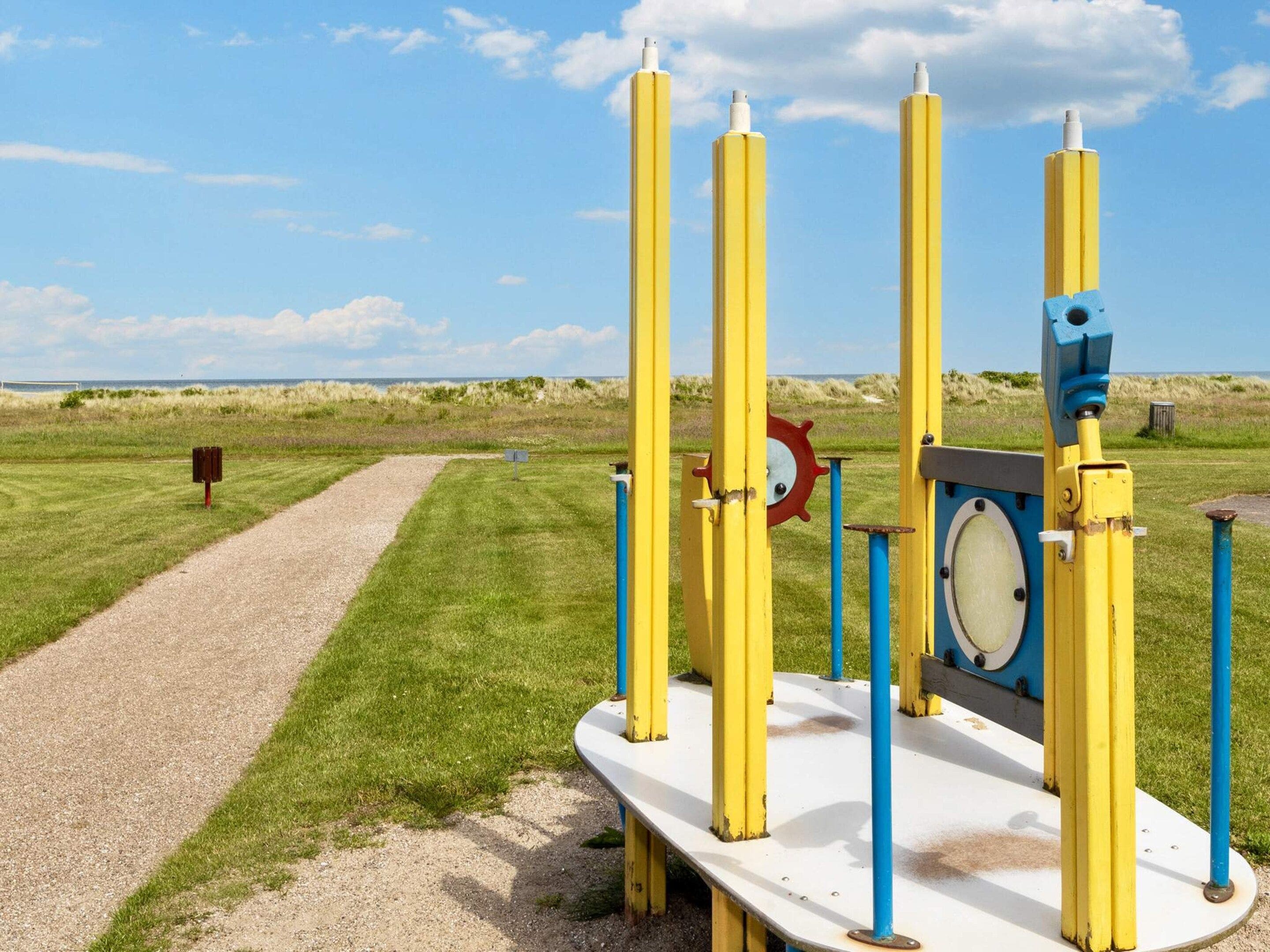 A playground with yellow and blue equipment, a gravel path, and a grassy field with a dune in the background.