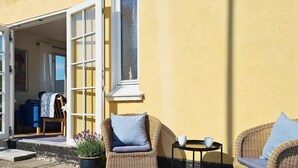 Ein Sitzbereich im Freien mit Korbstühlen, einem kleinen Tisch, einem Topfpflanzen und einem Gebäudeeingang mit Tür und Fenstern.