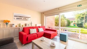 Un salon moderne avec un canapé rouge, un fauteuil gris et une table basse en bois. Une fenêtre offre une vue sur un jardin et un bâtiment à l’extérieur.