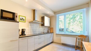 Une cuisine moderne avec des meubles blancs, un micro-ondes noir, une hotte en acier inoxydable et une fenêtre équipée de stores.