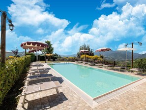 Une piscine entourée de chaises longues et de parasols, bordée de dalles de pierre et de verdure, avec des montagnes en toile de fond.