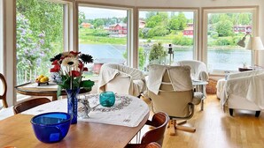 A dining area with a wooden table, chairs, and a vase with flowers. There are windows with a view of a lake and houses. - 4 Star Holiday Home in Vaddo (Väddö)