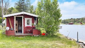 A small cabin with a porch, a table, and chairs, situated by a lake with a dock and a red building in the background. - 4 Star Holiday Home in Vaddo (Väddö)