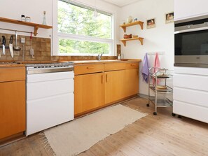 A kitchen with wooden cabinets, a stainless steel dishwasher, a window with a view of trees, and a wall-mounted oven. - 6 Person Holiday Home in Hadsund (Hadsund)