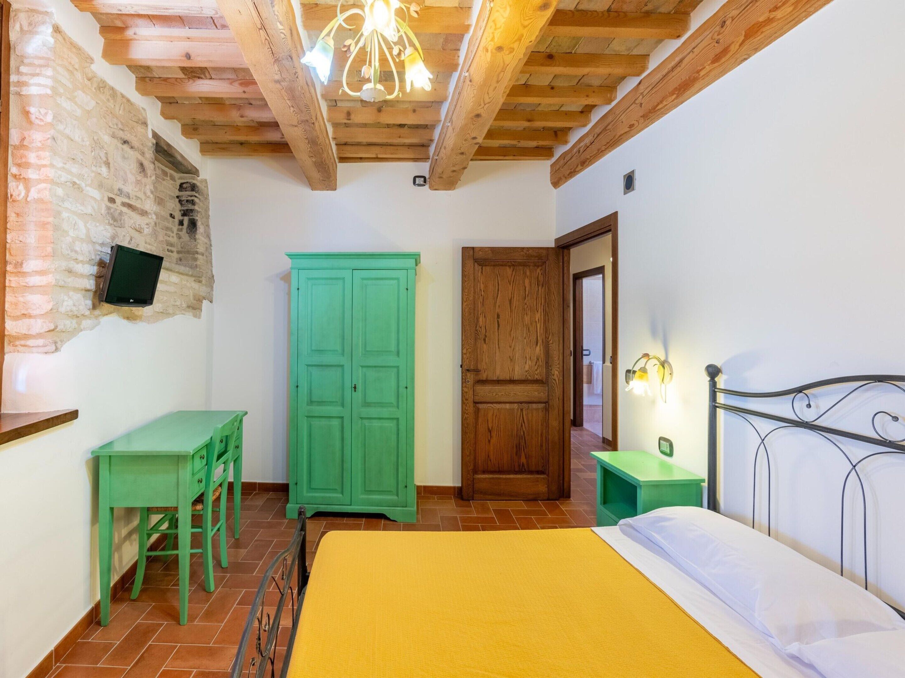 A bedroom with a bed, a green desk with chairs, a green wardrobe, a wooden door, and a ceiling light fixture.