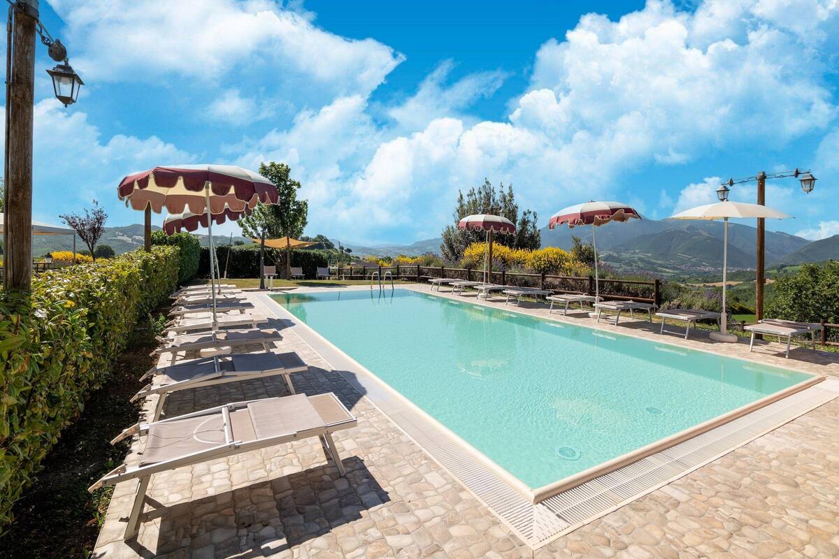A swimming pool with lounge chairs and umbrellas, surrounded by stone tiles and greenery, with mountains in the background.