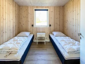 Una habitación pequeña con paneles de madera, dos camas individuales, una mesita blanca y una ventana que deja entrar luz natural.