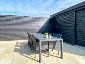 Un patio al aire libre con una mesa y sillas, piso de baldosas y pared de madera oscura.