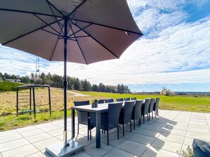 Un patio al aire libre con una mesa y sillas, un paraguas grande y vistas a un campo verde y árboles distantes.