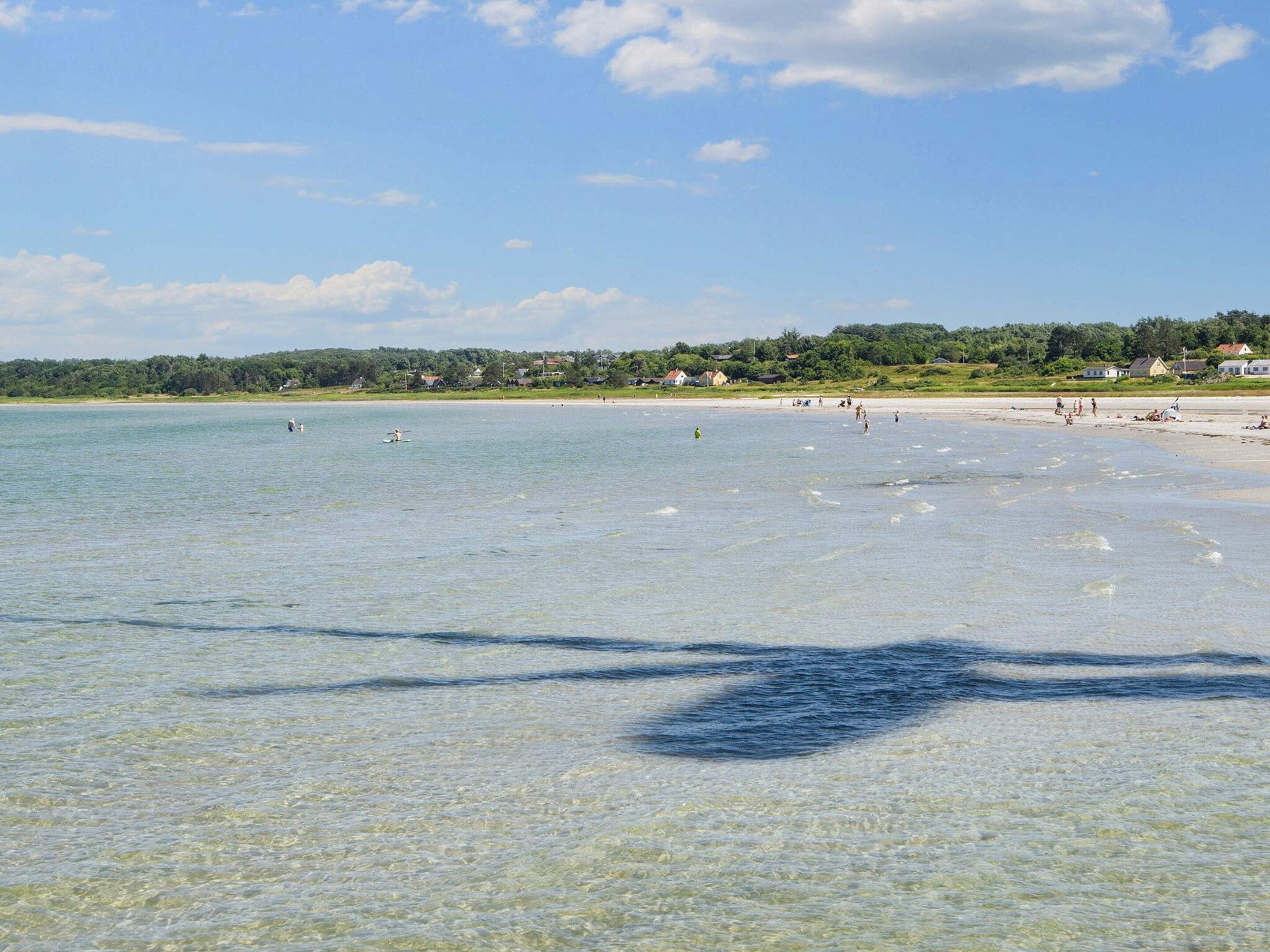 A beach with clear shallow water, a sandy shore, and a distant shoreline with greenery and buildings.