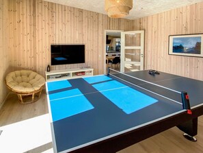 Una habitación con mesa de ping pong, televisor, una silla de mimbre y una puerta que da a otra estancia.