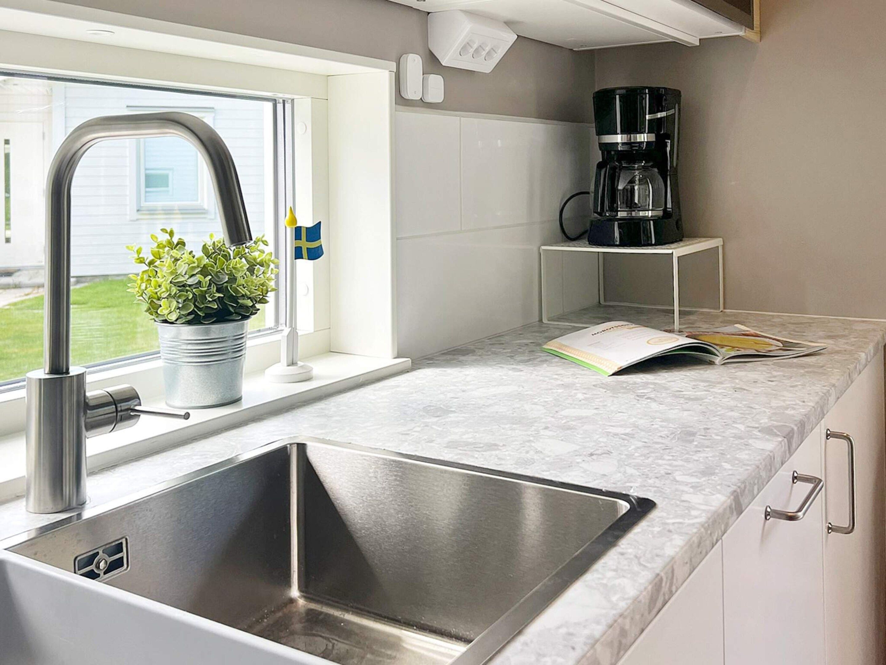 A kitchen with a stainless steel sink, a window with a flag, a potted plant, and a coffee maker on a shelf.