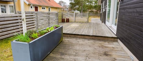 A wooden deck with a planter box containing plants, a fence, and a building with a window.