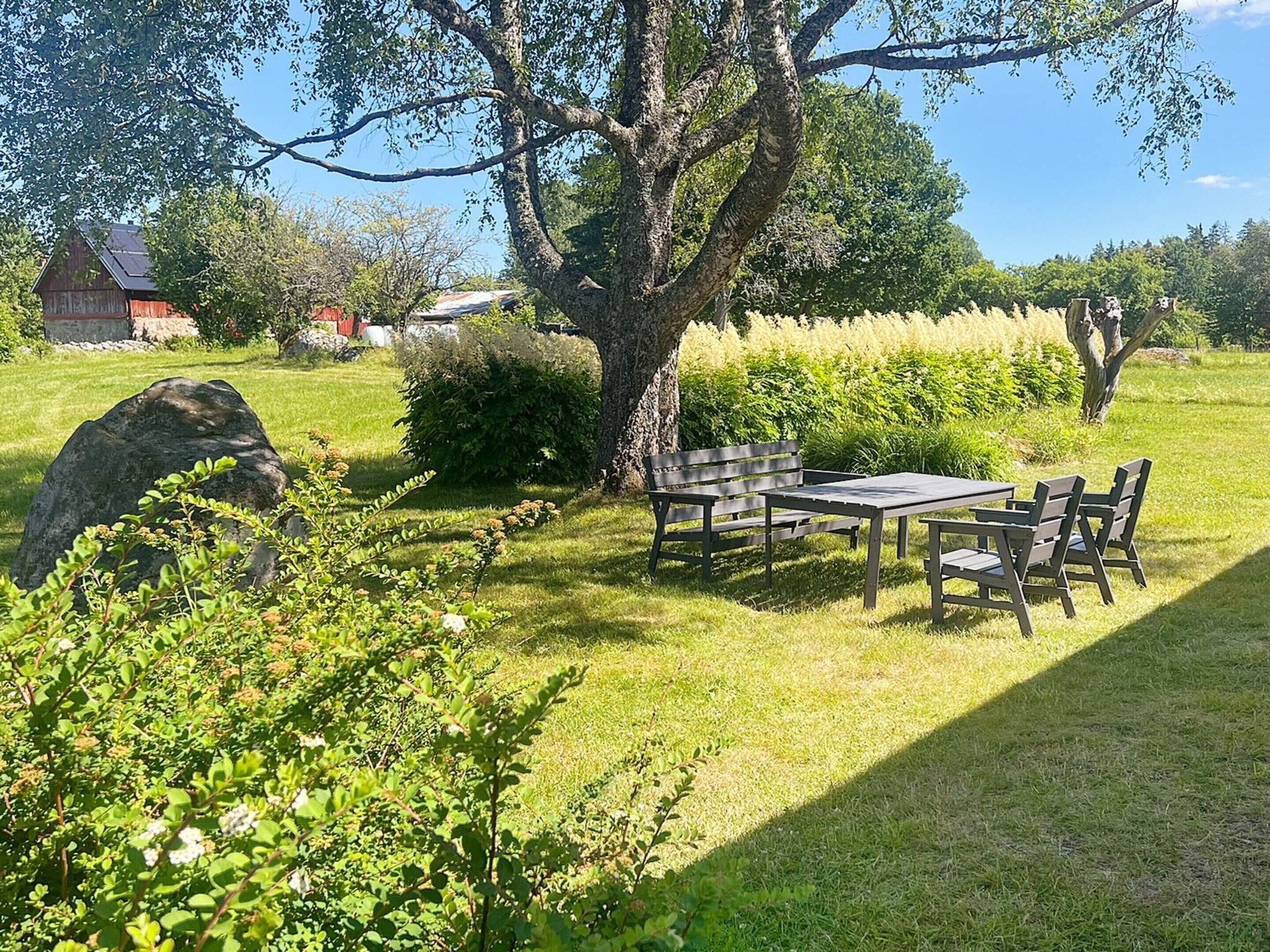 A grassy outdoor area with a table and chairs, a large tree, and a stone structure in the background.