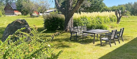 A grassy outdoor area with a wooden table and chairs, a large tree, and a stone structure in the background.