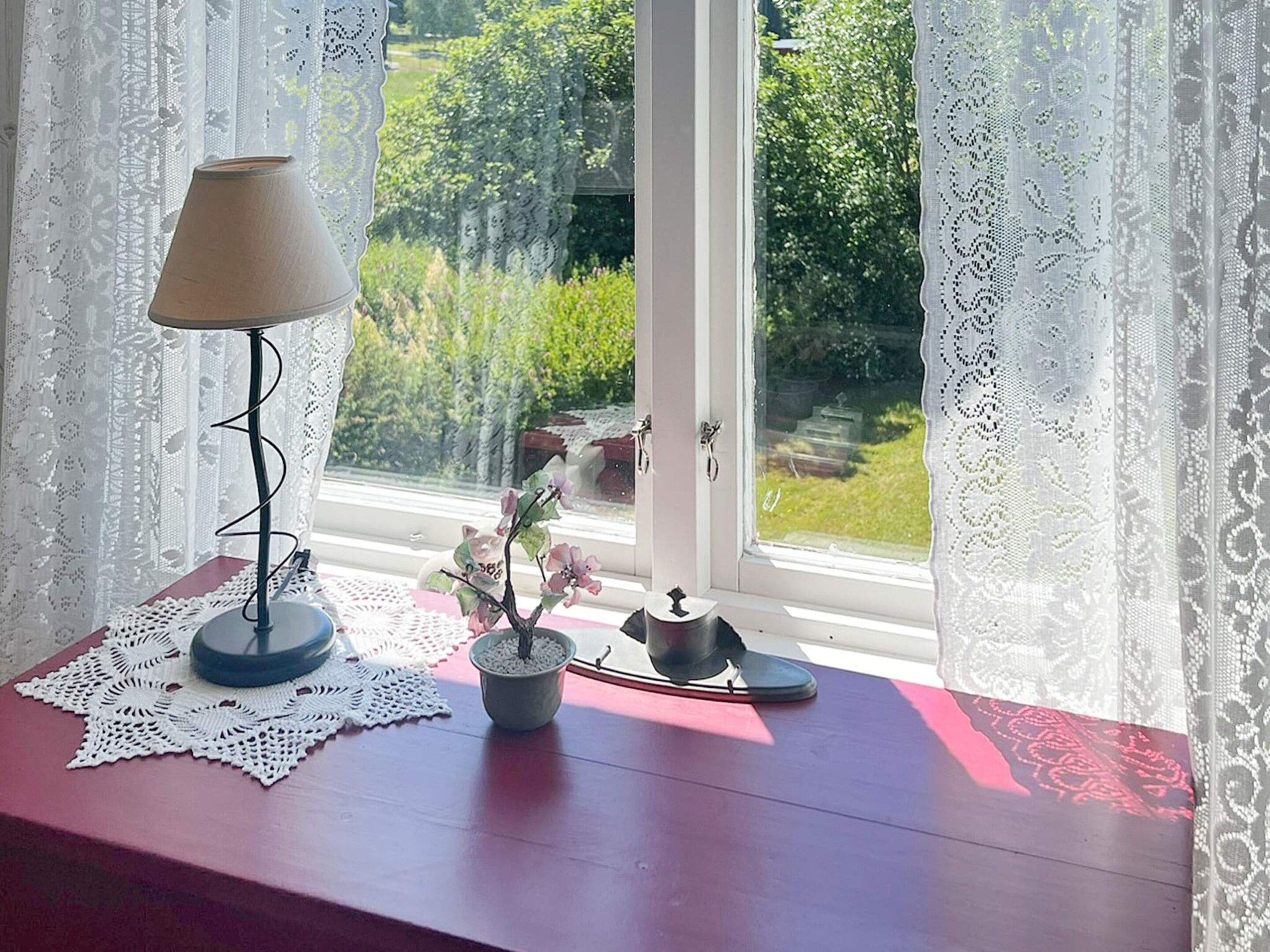 Una ventana con cortinas de encaje, una mesa con lámpara, una pequeña planta en maceta y un objeto decorativo.