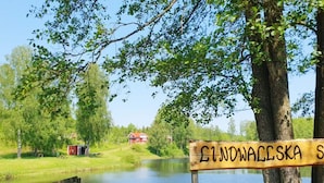 A wooden sign with the text "ŁODOWAŁEŚKA" in front of a calm lake, surrounded by trees and a small red building in the background. - 4 Star Holiday Home in Karlskrona (Karlskrona)