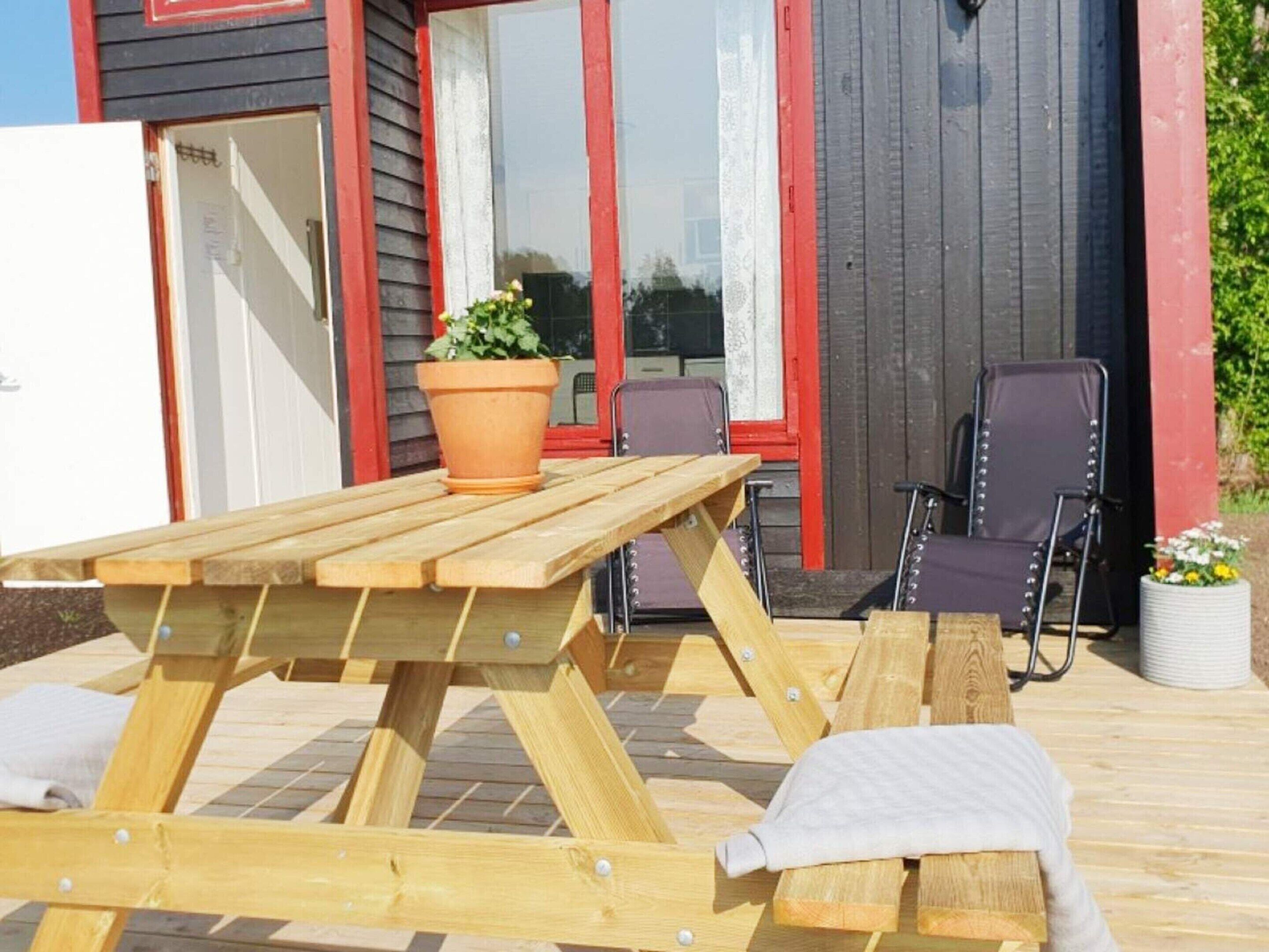 An outdoor wooden deck with a picnic table and chairs, a potted plant, and a building with red-framed windows in the background.