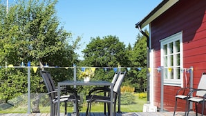 A wooden deck with a table and chairs, a red building with a window, and a garden in the background. - 5 Person Holiday Home in Hedekas (Hedekas)