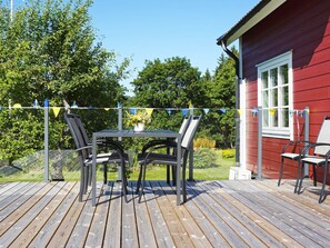 A wooden deck with a table and chairs, a red building with a window, and a garden in the background. - 5 Person Holiday Home in Hedekas (Hedekas)