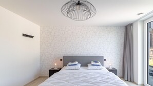 Une chambre moderne avec un grand lit, un luminaire suspendu original et une vue sur un immeuble par la fenêtre.