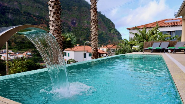 A swimming pool with a waterfall feature, surrounded by palm trees and a mountain backdrop.