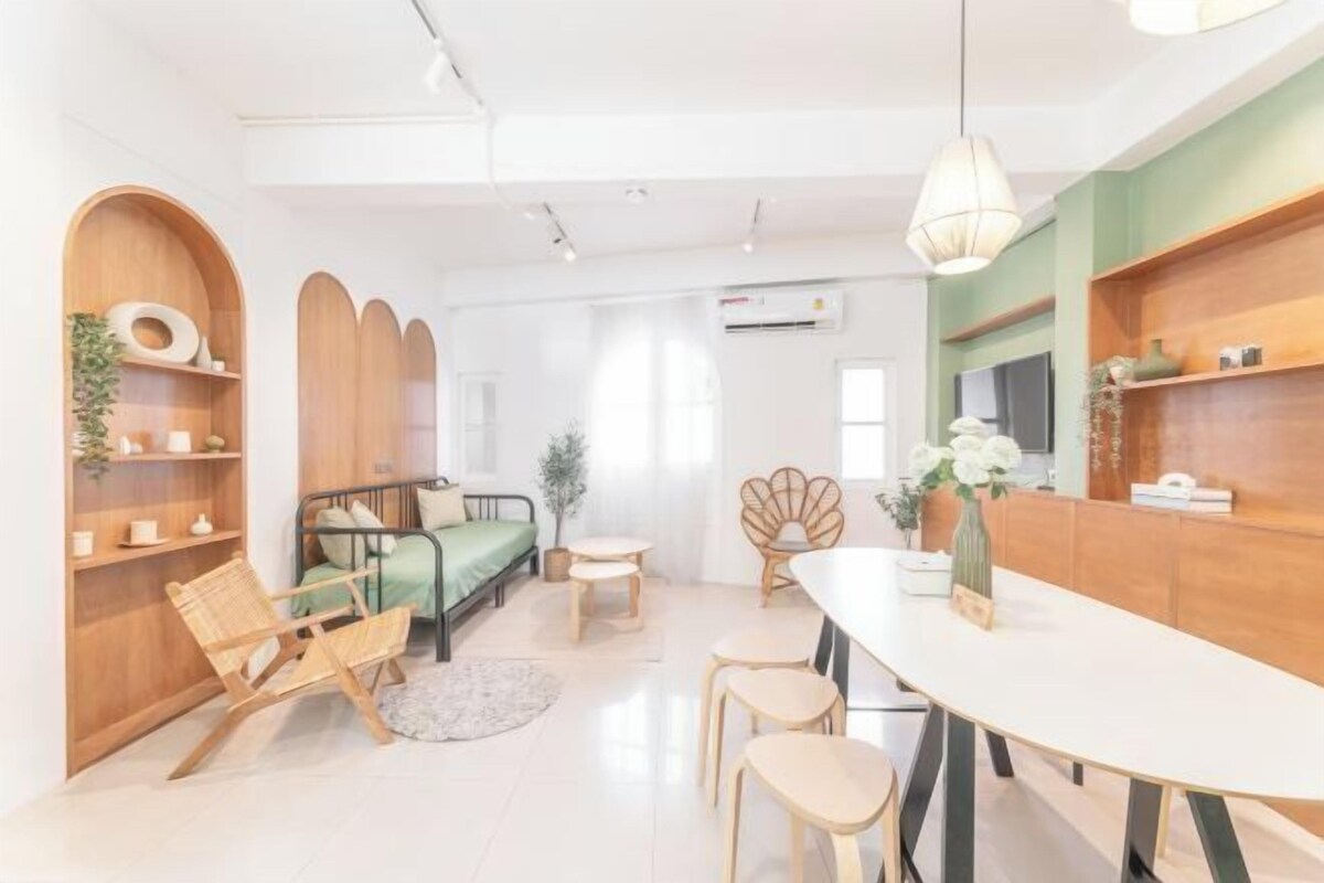 Una sala de estar moderna con un sofá, una mesa de comedor y sillas. Hay estantes con objetos decorativos y una planta.