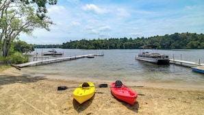 Una playa arenosa con dos kayaks, un muelle y un barco en un lago rodeado de árboles.