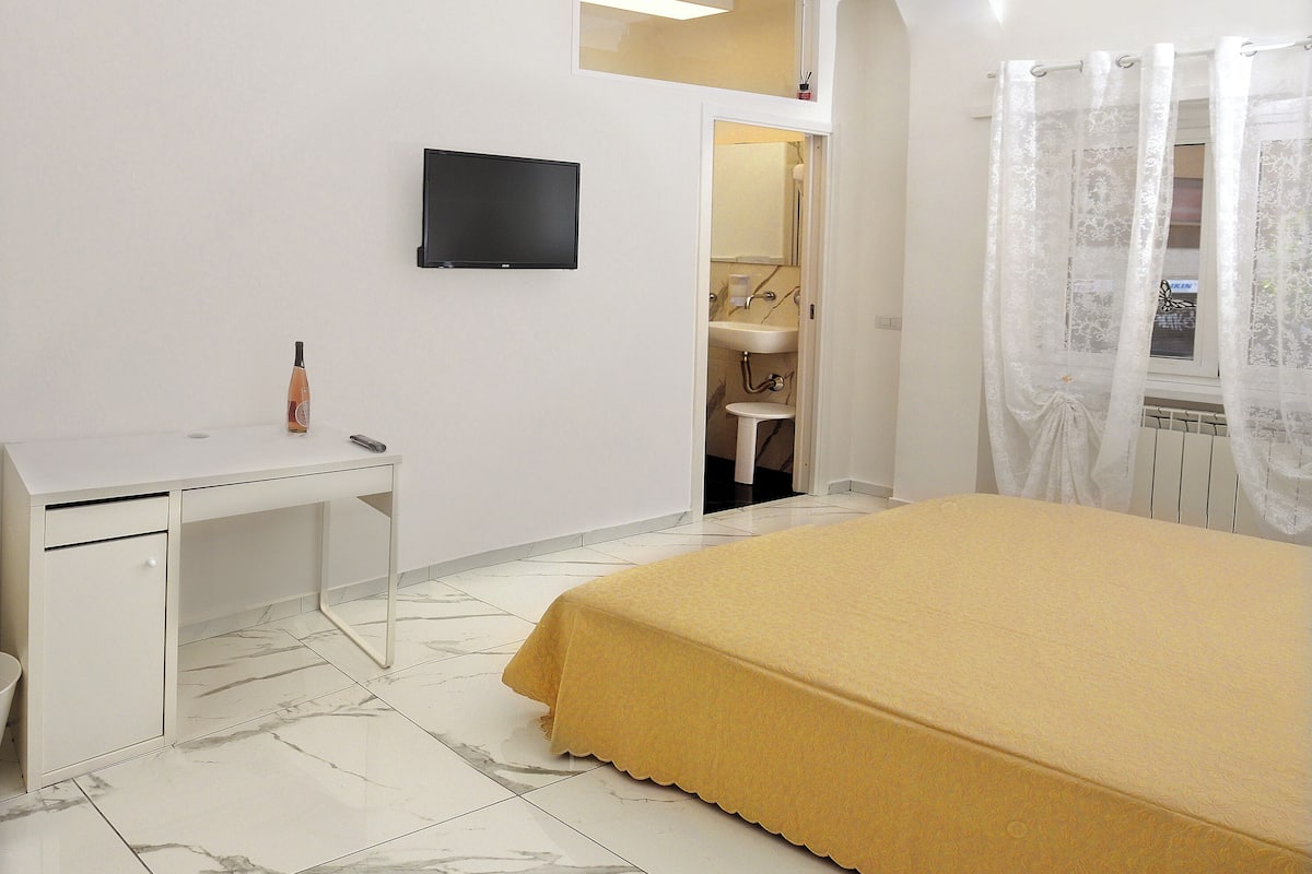 Un dormitorio con una cama, un escritorio, un televisor y un baño visible a través de una puerta abierta.