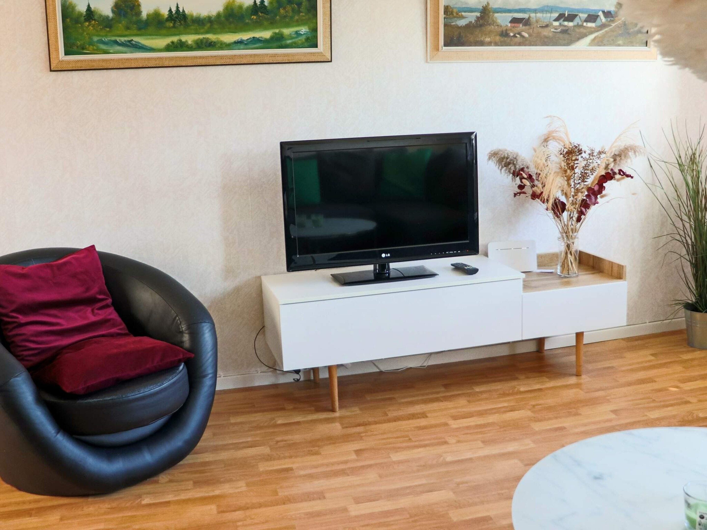 En stue med en sort læderstol, et hvidt TV-møbel med et fladskærms TV, en fjernbetjening og en vase med tørrede planter.